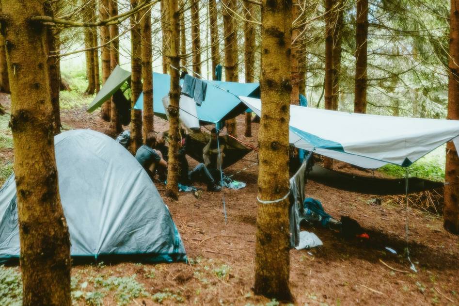 Person building a survival shelter in forest with fire nearby and gear laid out